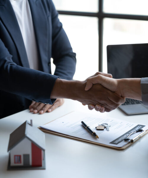 Two young Asian businessmen shake hands after signing a contract to invest in a village project. real estate, with businesswomen joining in showing joy and clapping in the office.